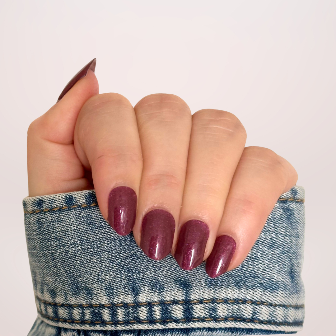 Close-up van een hand met donkerpaarse gellakstickers, versierd met subtiele glitter, die een luxe en sprankelende uitstraling geven voor een elegante nagellook