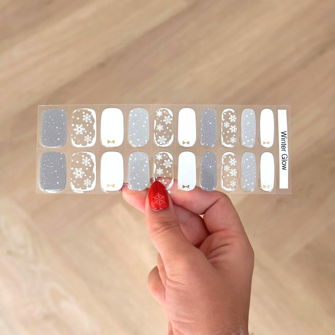 Winters gellaksticker design met elke nagel een ander design. De ene nagel een witte basis met subtiele glitter en een kleine gouden strik, een andere nagel is een grijze basis met glitter met subtiele witte stippen en een andere nagel is als basis transparant met witte opvulling en sneeuwvlokken