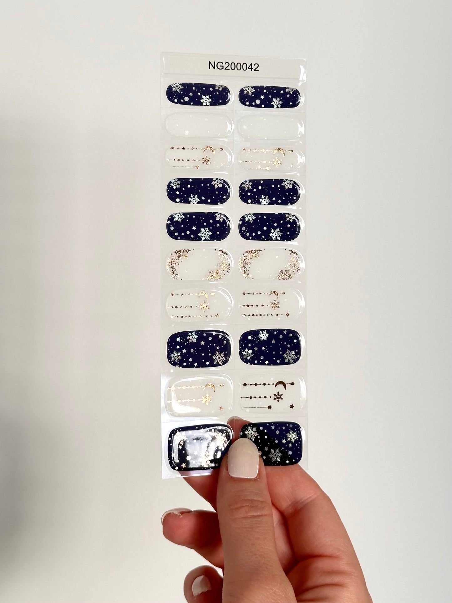 Winterse nail art. De ene nagel heeft een witte achtergrond met gouden details zoals kleine sneeuwvlokjes en maantjes, de andere nagel heeft als ondergrond donker blauw met witte sneeuwvlokken en sneeuw stippen. Perfect voor de winter