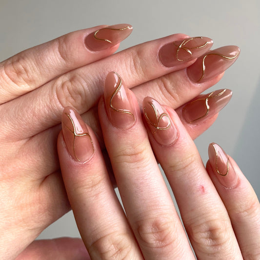 Lange almondvormige nagels met een nude basis en subtiele gouden swirl lijnen. 