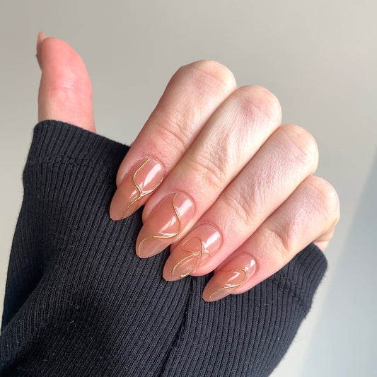 Lange almondvormige nagels met een nude basis en subtiele gouden swirl lijnen. 