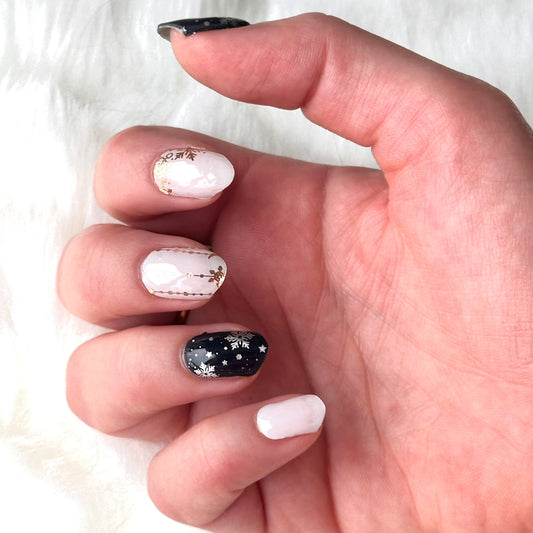 Winterse nail art. De ene nagel heeft een witte achtergrond met gouden details zoals kleine sneeuwvlokjes en maantjes, de andere nagel heeft als ondergrond donker blauw met witte sneeuwvlokken en sneeuw stippen. Perfect voor de winter